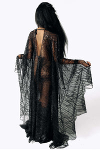 Back view of Cameron wearing the Azrael Black Winged Sleeve Maxi Dress, a full-length black mesh dress from SCATHED's Fall Winter 2025 Reaper collection while standing with her arms extended from her waist, showing the draping bat winged sleeves.