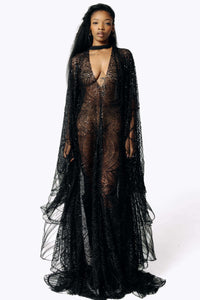 Frontal view of Cameron wearing the Azrael Black Winged Sleeve Maxi Dress, a full-length black mesh dress from SCATHED's Fall Winter 2025 Reaper collection while standing with her arms at her sides, showing the layering of the bat winged sleeves.