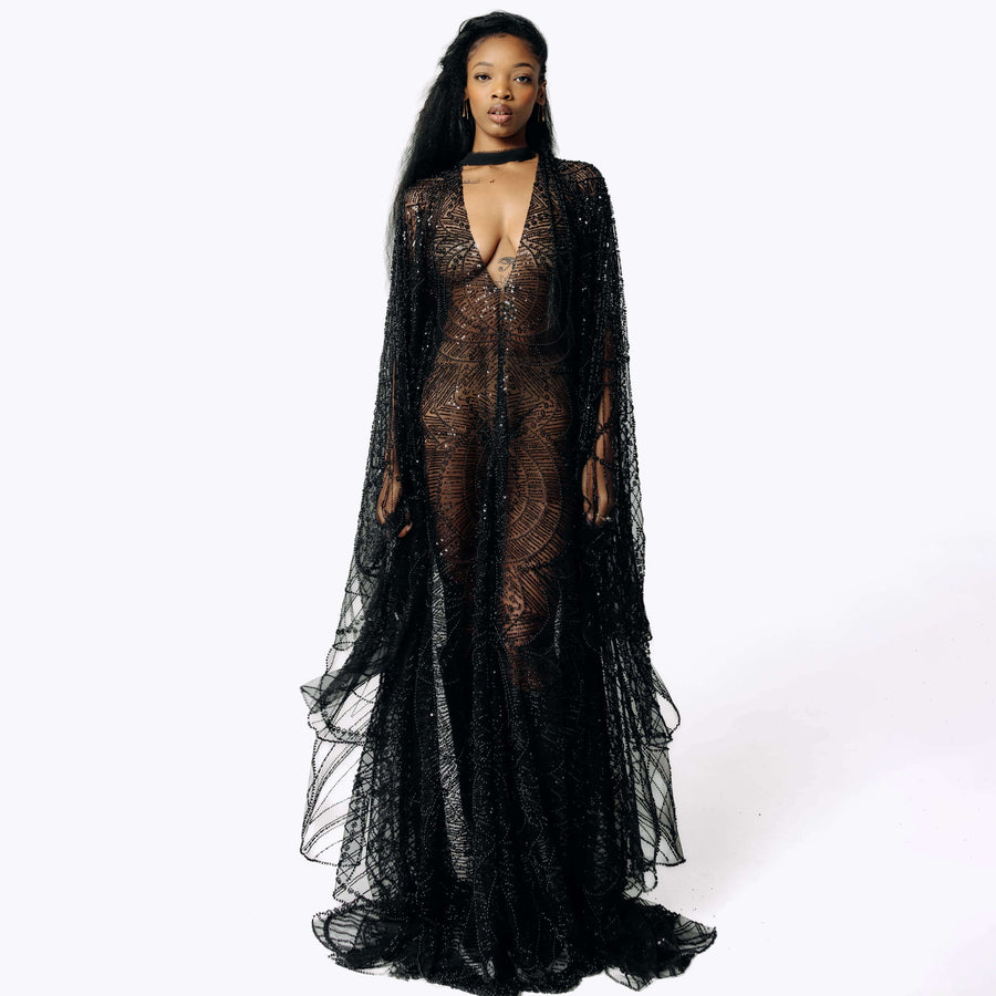 Frontal view of Cameron wearing the Azrael Black Winged Sleeve Maxi Dress, a full-length black mesh dress from SCATHED's Fall Winter 2025 Reaper collection while standing with her arms at her sides, showing the layering of the bat winged sleeves.