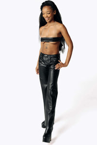 Angled view of Cameron wearing the Dicentra Leather Bralette with an exposed midriff and the Pale Rider Cactus Leather Pants from SCATHED's Fall Winter 2025 Reaper collection while staring at the white floor smiling. Cameron is also wearing black high-heeled platform leather boots. 