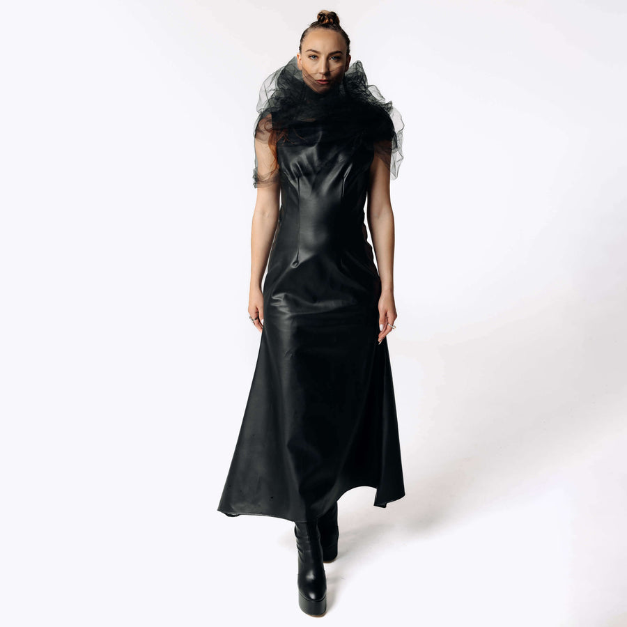 Front view of Caitlin wearing the Kalma Cactus Leather Maxi Dress, a full-length black leather dress that flares out from the hips with the Wraith Black Tulle Collar from SCATHED's Fall Winter 2025 Reaper collection. Caitlin also wears high-heeled black platformed ankle boots.