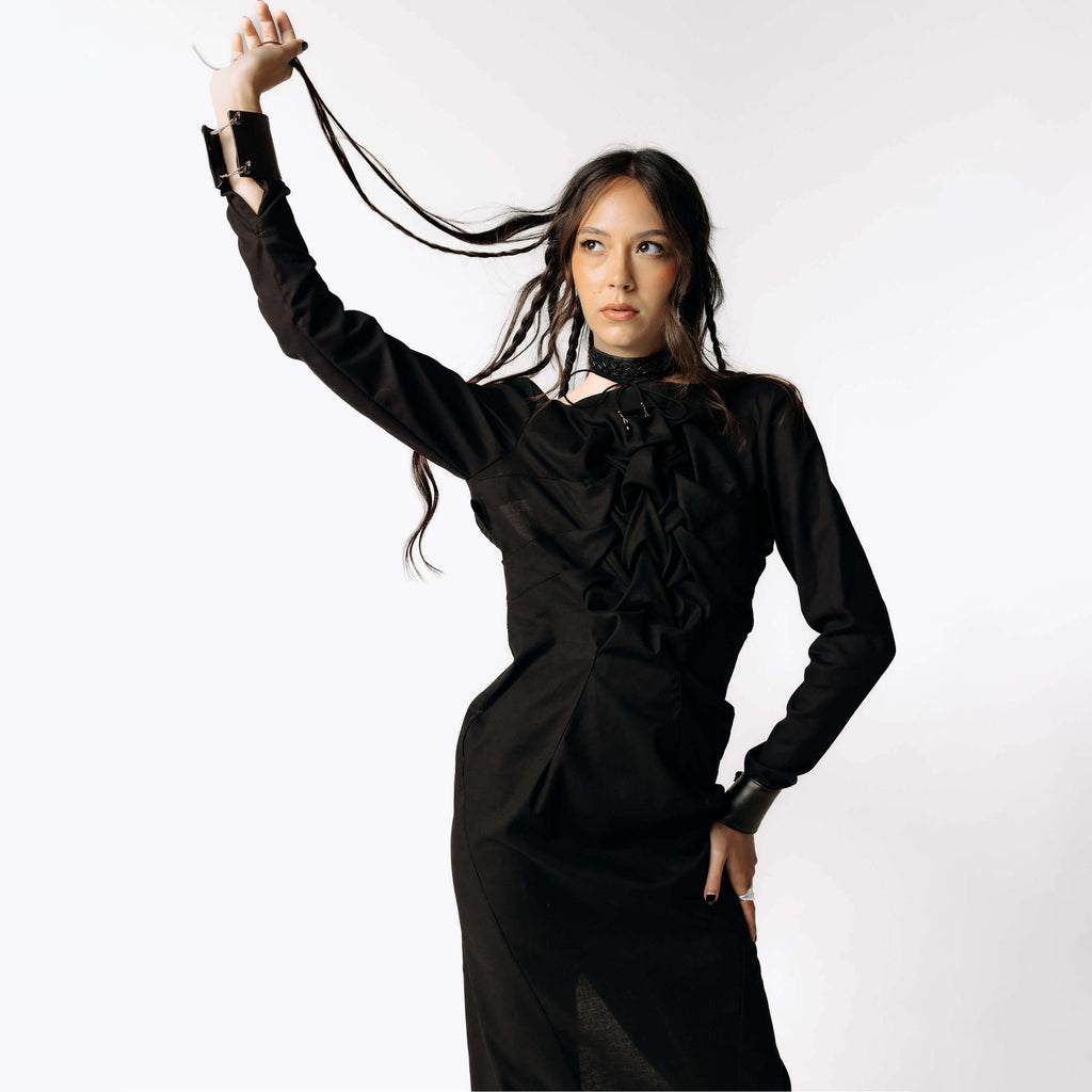 Frontal view of Eliza wearing the Mors Black Cotton Dress with ruffling in the chest from SCATHED's Fall Winter 2025 Reaper collection, while playing with her hair and looking off to the left.