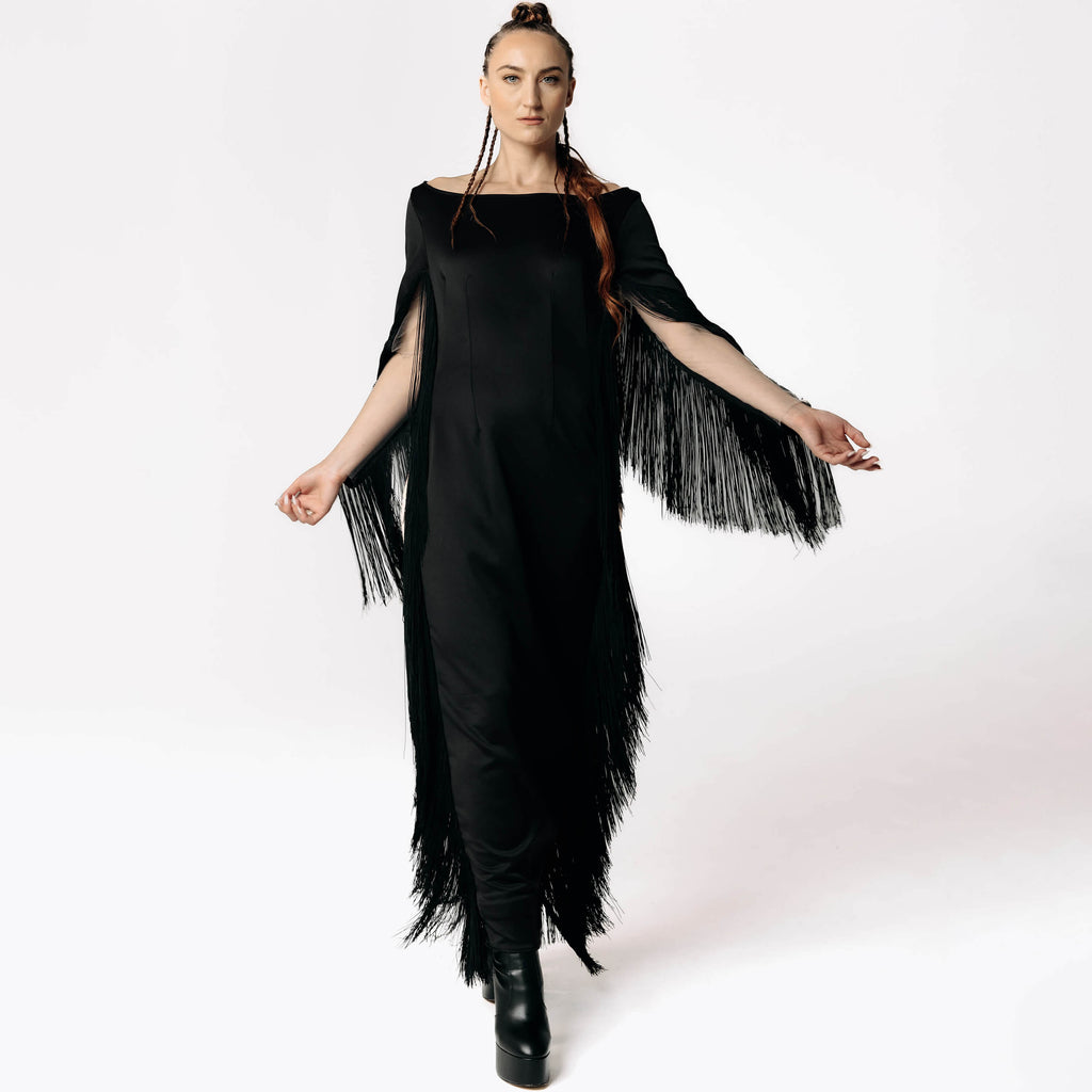 Frontal view of Caitlin stepping towards camera while wearing the Revenant Black Dress with fringed sleeves and a high thigh-slit from SCATHED's Fall Winter 2025 Reaper collection.