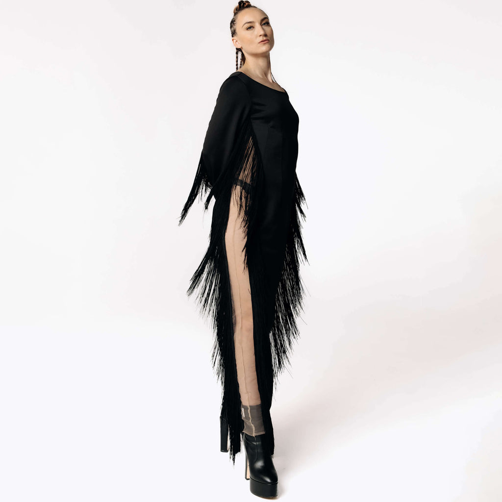 Side view of Caitlin stepping towards camera while wearing the Revenant Black Dress with fringed sleeves and a high thigh-slit from SCATHED's Fall Winter 2025 Reaper collection.