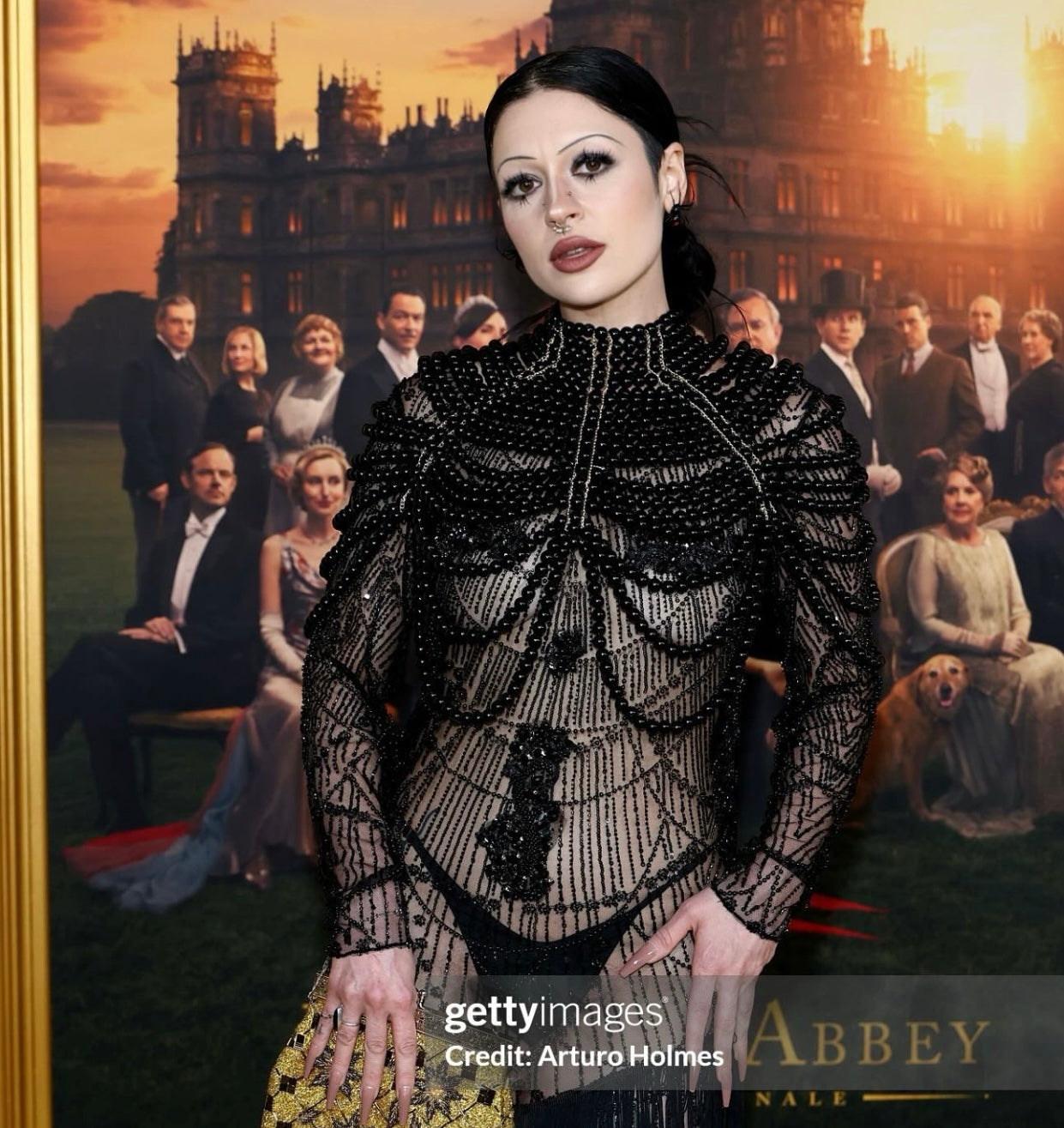 Samantha Lubrano wears the Morta Beaded Gown for SCATHED's F/W25 Reaper Collection to the Downtown Abbey premiere event while standing in front of a poster depicting scenes from the show. Image Credit: Arturo Holmes, Getty Images