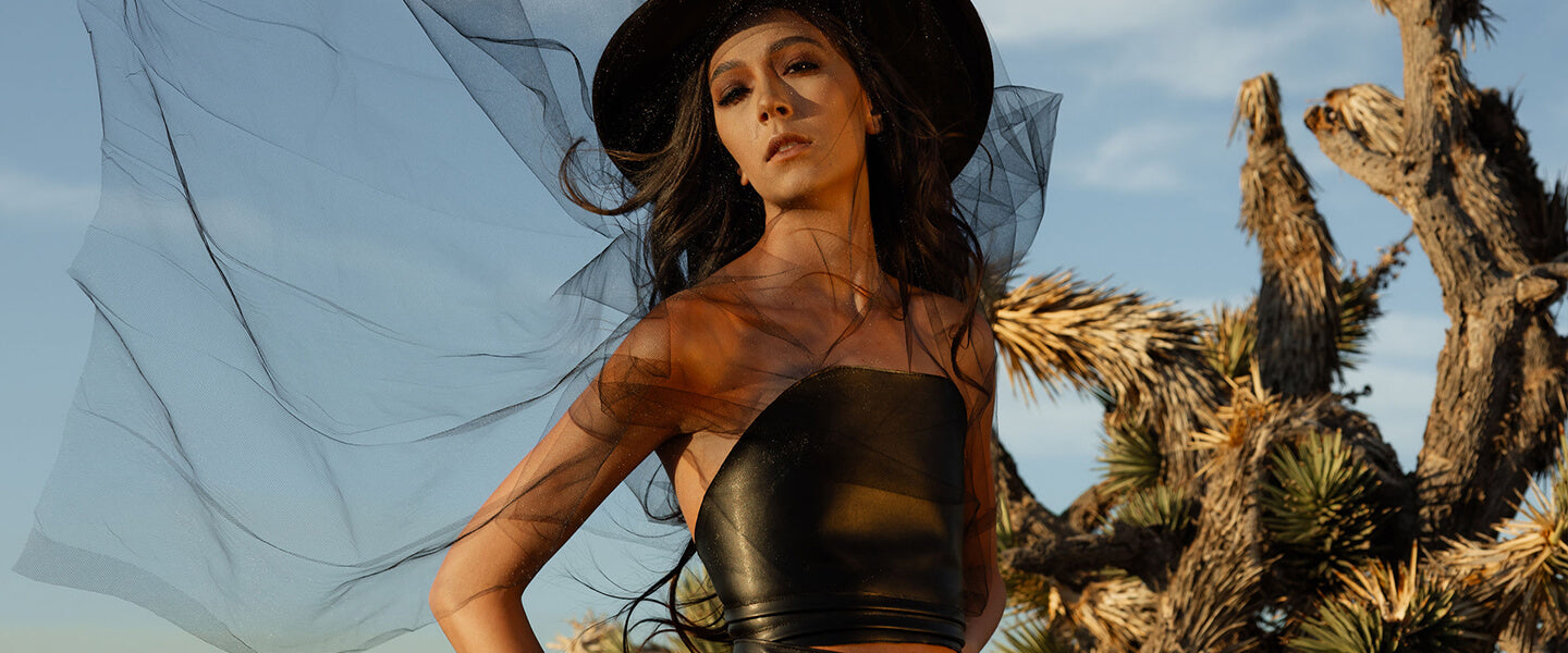 Woman in a black outfit and hat with a flowing fabric against a desert landscape. Discover gothic-inspired leather skirts from SCATHED>