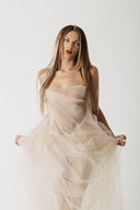 A woman modeling a strapless eco-tulle dress in a light champagne beige color, holding out the sides of the voluminous skirt and corset against a plain light gray studio background.