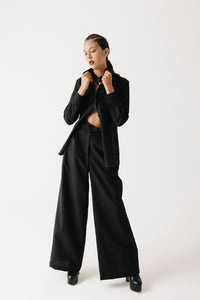 Model wearing the Mourner Shirt, a cropped black button-up in organic bamboo cotton, worn open over trousers