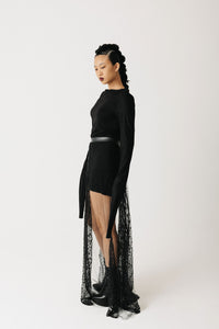 A model wearing a black long-sleeve mini dress with a high-low sheer lace overlay beaded skirt, styled with a black belt, against a plain light gray studio background.