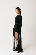 A model wearing a black long-sleeve mini dress with a high-low sheer lace overlay beaded skirt, styled with a black belt, against a plain light gray studio background.