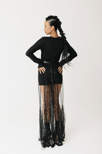 A model is shown from the back wearing a black long-sleeve top and a sheer black beaded maxi skirt with a wide sheer overlay, set against a plain light gray studio background.