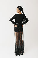 A model is shown from the back wearing a black long-sleeve top and a sheer black beaded maxi skirt with a wide sheer overlay, set against a plain light gray studio background.