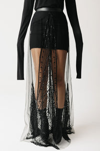 A model wearing a black long-sleeve top and a black mini skirt with a dramatic sheer black eco-tulle overlay beaded skirt featuring intricate lace and beadwork details.