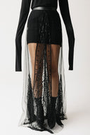 A model wearing a black long-sleeve top and a black mini skirt with a dramatic sheer black eco-tulle overlay beaded skirt featuring intricate lace and beadwork details.