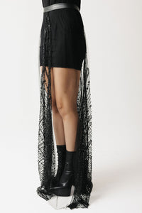 A model's lower body wearing a black mini dress with a long, sheer black beaded eco-tulle overlay skirt and black platform ankle boots, photographed against a plain white studio background.
