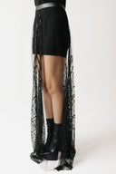 A model's lower body wearing a black mini dress with a long, sheer black beaded eco-tulle overlay skirt and black platform ankle boots, photographed against a plain white studio background.