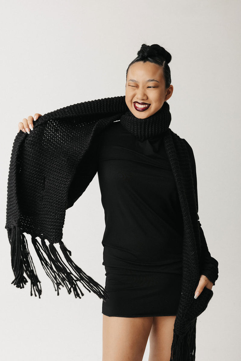 A woman modeling a chunky black knit wrap scarf with fringe and pockets, worn over a black turtleneck dress against a light gray studio background.