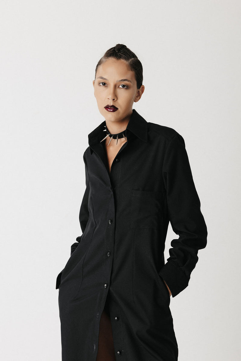 A model with dark hair pulled back wears the widow long black button-up shirt dress with a collar and front pockets, styled with a black choker against a light gray studio background.