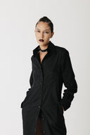 A model with dark hair pulled back wears a long black button-up shirt dress with a collar and front pockets, styled with a black cactus leather choker with spiked studs against a light gray studio background.