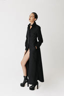 A model wearing the widow long black shirt dress with a high slit, styled with black platform ankle boots, posing against a plain white studio background. side view