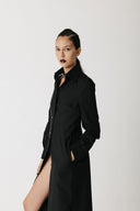 A model wearing a long black button-up shirt dress with a high side slit, styled with a black choker, against a plain light gray studio background.