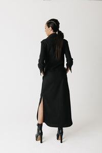 A woman is shown from behind wearing the widow long black shirt dress with a high collar and a thigh-high slit, paired with black ankle boots, against a plain white studio background.