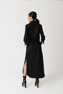 A woman is shown from behind wearing the widow long black shirt dress with a high collar and a thigh-high slit, paired with black ankle boots, against a plain white studio background.