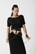 A model wearing a black short-sleeve crop top with a cross-shaped button detail on the chest, paired with a black skirt featuring side cutouts and gold chain draping, against a plain light gray background.