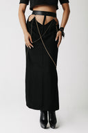 A model wearing the penance black midi skirt with a cutout back, a black leather belt, and a draped gold chain detail, paired with black boots against a light gray background.