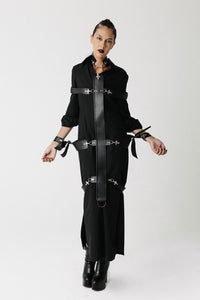 A model wearing a black long-sleeve shirt dress, with the stuart cactus leather full body harness, photographed against a plain white studio background.