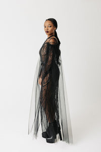 A model wearing a sheer black lace and tulle dress with fringe details, standing in profile against a plain white studio background.