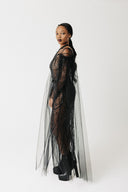A model wearing a sheer black lace and tulle dress with fringe details, standing in profile against a plain white studio background.