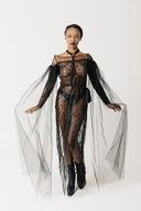 A model wearing a sheer black beaded tulle and lace off-the-shoulder dress with fringe details and long sheer eco-tulle sleeves, paired with black boots, against a plain white studio background.