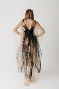 A woman shown from the back wearing a sheer layered tulle gown in nude and black, photographed against a plain light background.