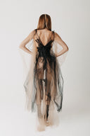 A woman shown from the back wearing a sheer layered tulle gown in nude and black, photographed against a plain light background.