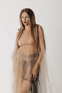A model with long brown hair wearing a sheer beige eco- tulle gown over black underwear, posing against a plain white studio background.