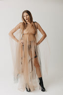 A woman modeling a sheer beige eco-tulle dress paired with black high-heeled boots, against a plain light gray studio background.