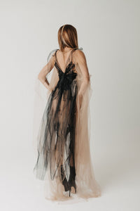 A woman stands with her back to the camera wearing a sheer black lace dress layered with a long, flowing nude-beige tulle cape on a plain light gray studio background.