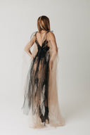 A woman stands with her back to the camera wearing a sheer black lace dress layered with a long, flowing nude-beige tulle cape on a plain light gray studio background.