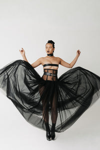 A model wearing a black strapless bandeau top, a matching black waist belt with a silver buckle, and a voluminous black tulle skirt, posing with her arms outstretched against a plain white studio background.