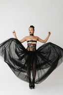 A model wearing a black strapless bandeau top, a matching black waist belt with a silver buckle, and a voluminous black tulle skirt, posing with her arms outstretched against a plain white studio background.