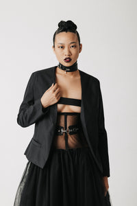 A model wearing a black structured bamboo cotton blazer over a sheer black strappy top and black skirt, accessorized with a black choker, against a plain light gray studio background.