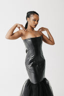 A model wearing a strapless black cactus leather corset dress with a tulle mermaid skirt, posing against a light gray studio background.