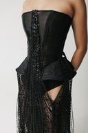 A close-up of a model wearing a black strapless corset-style dress with sheer mesh panels, structured black fabric, and long hanging hand-=beaded fringe.