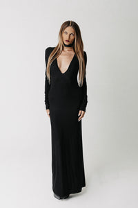 A woman modeling a long black dress with long sleeves, a deep plunging neckline, and a floor-length silhouette, photographed against a plain light gray studio background.