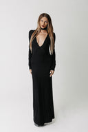 A woman modeling a long black dress with long sleeves, a deep plunging neckline, and a floor-length silhouette, photographed against a plain light gray studio background.