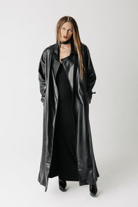 A model wearing a long black cactus leather trench coat over a black dress, standing against a light gray studio background.
