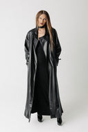 A model wearing a long black cactus leather trench coat over a black dress, standing against a light gray studio background.