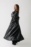 A woman modeling a long, glossy black cactus leather trench coat with a high collar and waist tie, photographed against a plain light gray studio background.