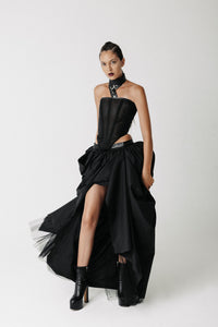 A model wearing a black strapless corset with a high-low draped Sisi skirt, and black platform ankle boots, posed against a plain light gray studio background.
